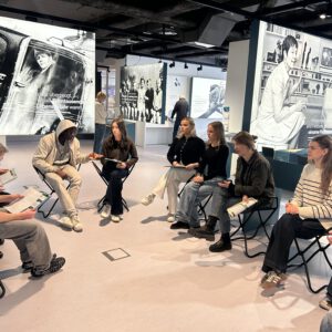 Schüler sitzen in einer Diskussionsrunde im Kreis. Im Hintergrund sind Plakate einer Ausstellung zu sehen.